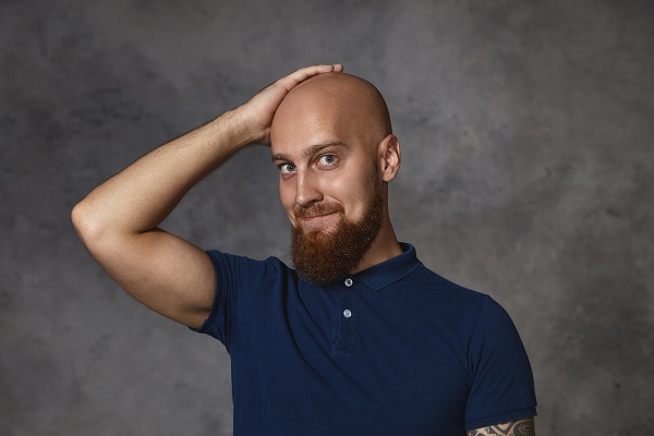 bald man with one hand on his head looking at the camera with a faint smile
